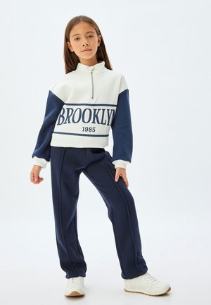 Sweat à coloris bloc blanc et marine avec demi-fermeture éclair, arborant l'inscription "BROOKLYN 1985" sur le devant, associé à un pantalon de survêtement marine.