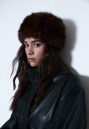 Eine Frau mit langen, dunklen Haaren trägt einen dunkelbraunen Pelzhut und eine schwarze Lederjacke, während sie gegen einen schlichten hellen Hintergrund zur Kamera blickt.