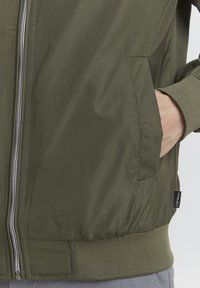 La veste bomber olive possède un tissu léger et lisse, une fermeture éclair à l'avant, des poignets élastiques et des poches latérales. Détails texturés subtils.