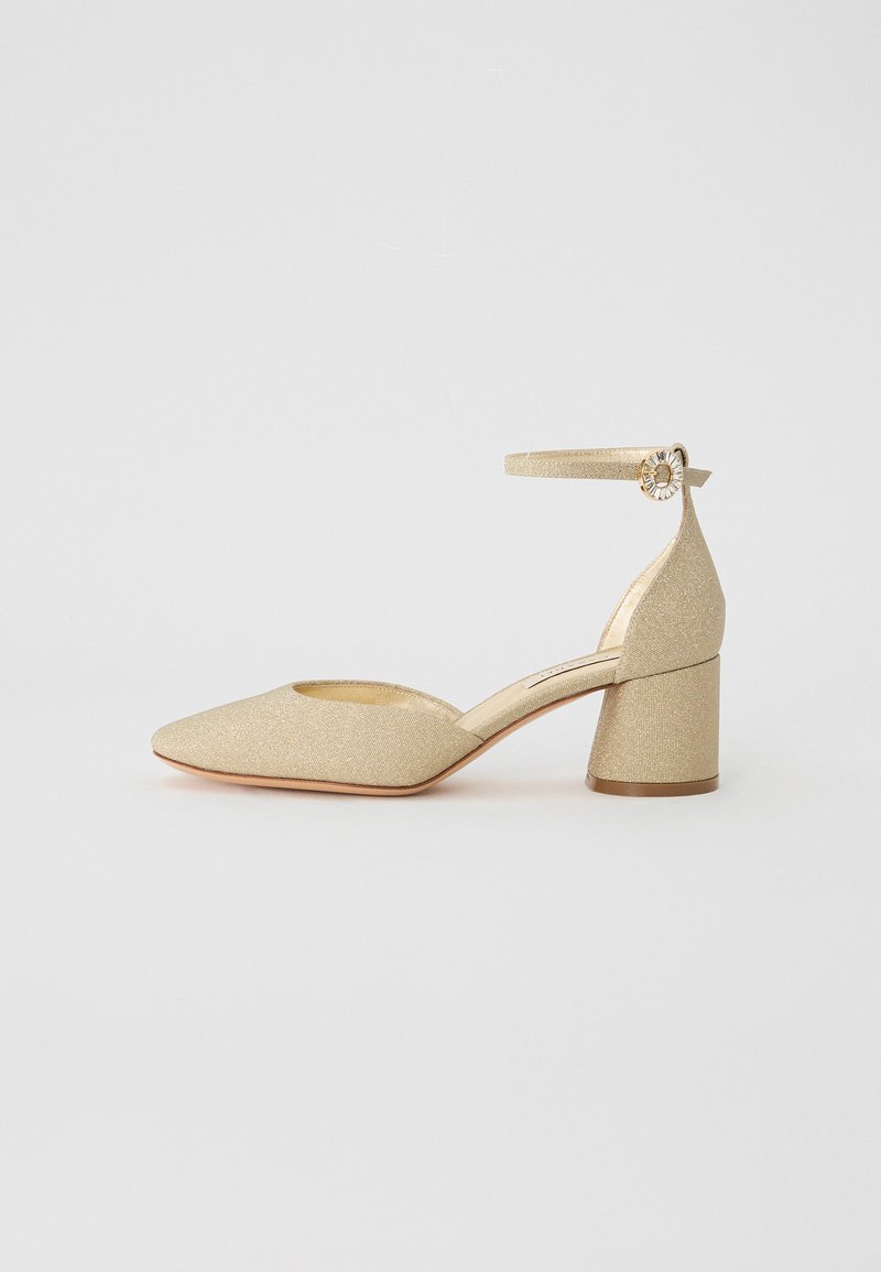 Chaussure basse à talon doré pailleté avec un bout pointu, une lanière à la cheville et des éléments décoratifs. Texture lisse avec une finition brillante.