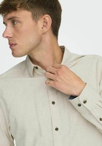Camisa de botón de color beige claro con un bolsillo en el pecho, textura visible y botones oscuros. El puño presenta un acento azul y un diseño ajustado.