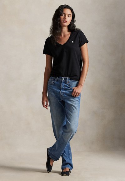 Γυναίκα με κυματιστά μαλλιά όρθια, που φοράει μαύρο T-shirt με V-λαιμόκοψη και λογότυπο, μπλε τζιν και μαύρα παπούτσια slip-on, σε απλό φόντο.