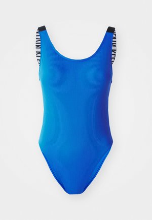 Sinine ribadega ühte tükki ujumistrikoo kõrge lõikega sääre, ümar dekoltee ja musta-valgete aktsentidega mustrid pannalrihmadel.