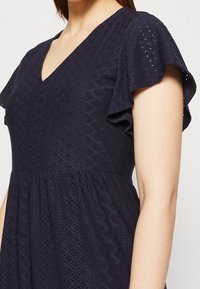 Eine Frau trägt ein dunkelblaues Kleid mit kurzen schwingenden Ärmeln und einem strukturierten geometrischen Spitzenmuster, das über einen V-Ausschnitt verfügt.