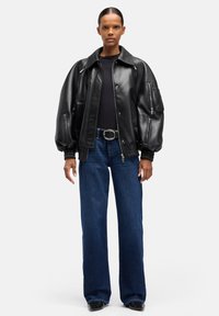 Czarna skórzana kurtka bomber o luźnym kroju, z ściągaczami na mankietach, dwoma kieszeniami na suwak, noszona na czarnej bluzce i zestawiona z niebieskimi szerokimi spodniami jeansowymi.