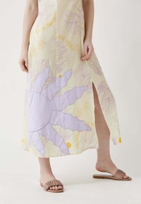 Maxi-mekko keltaisesta kankaasta, jossa on violetti auringon grafiikka, halkiot sivuilla, ja kukkakuoseja sekä tekstiyksityiskohtia. Yhdistetty ruskeisiin liukukenkäisiin.