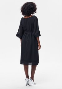 Kobieta o kręconych włosach, ubraną w czarną sukienkę midi z rękawami do łokci oraz srebrne buty na wysokim obcasie, stojąca tyłem na białym tle.