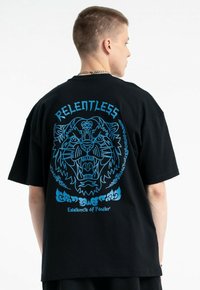 Czarny, oversize T-shirt z niebieską grafiką twarzy tygrysa, z napisem "RELENTLESS" powyżej i "Essence of Power" poniżej w dekoracyjnym stylu.