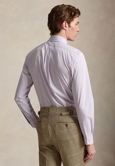 Muž nosi svetlofialovú prúžkovanú košeľu zastrčenú do hnedých nohavíc s vzorom kĺbovej labky, pozerajúci z profilu na jednoduchom pozadí.