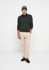 Uomo che indossa un maglione verde a zip fino al petto, una polo blu navy, pantaloni beige, mocassini marroni e un berretto a scacchi, in piedi contro uno sfondo bianco semplice.