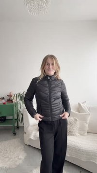 Sort quiltet jakke med stående krave og foran lynlås, med diagonale syningmønstre. Bæres over sorte bukser i et lyst rum.