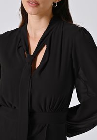 Schwarze Bluse mit einem Schlüssel-ausschnitt und gebundenem Detail. Hergestellt aus einem glatten, leichten Stoff, mit geraffter Taille und transparenten Ärmeln.