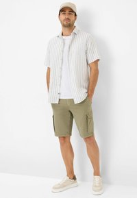 Camicia a maniche corte a righe in bianco e grigio chiaro, abbinata a pantaloni cargo verde oliva. T-shirt bianca sotto e sneaker beige.