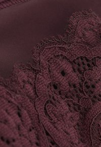Tissu bourgogne avec une dentelle complexe ornée de motifs floraux et de bords festonnés. Texture lisse avec des accents de broderie détaillés.