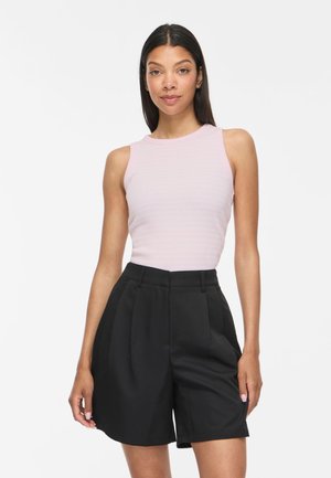 Femme portant un haut rose clair sans manches et un short noir taille haute sur mesure, debout devant un fond blanc uni.