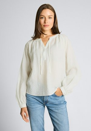 Femme portant un chemisier blanc texturé ample à manches longues et un jean bleu clair, debout avec une main dans la poche, devant un fond uni.