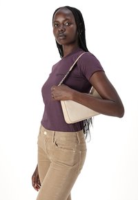 Beige shoulder bag with a structured design, worn with a fitted purple t-shirt and beige corduroy pants, featuring front pockets and buttons.