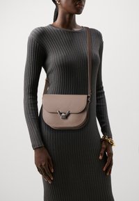 Lysebrun lædertaske med flaplukning og metallisk accent. Bæres over en struktureret mørkegrå ribbet kjole.