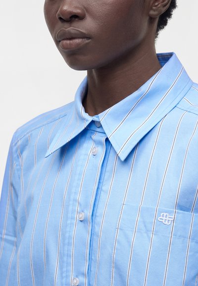 Gros plan d'une personne portant une chemise bleu clair à rayures verticales, boutonnée, avec un col pointu et un petit logo brodé sur la poche.