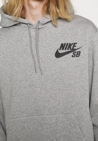Šedá mikina s kapucí na stažení, s černým logem Nike SB potisknutým na hrudi. Měkká, strukturovaná látka s klokaní kapsou.