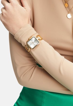 Montre carrée en or avec un cadran blanc, bracelet en métal maillé, portée sur un poignet nu. Haut à manches longues beige clair et tissu vert en dessous.