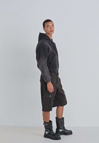 Zwarte rits hoodie met elastische mouwen, gecombineerd met cargo shorts tot de knie en kniehoge zwarte laarzen met gespen.