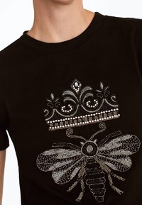 Sort bomulds T-shirt med detaljeret sølvbroderi af en krone over en bi. Designet inkluderer intrikate mønstre og teksturer.