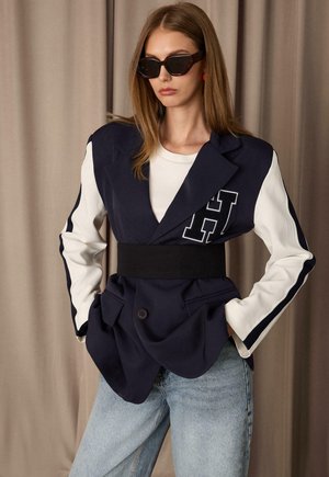 Femme portant un blazer oversize bleu marine et blanc avec une ceinture noire, de grandes lunettes de soleil noires, une chemise blanche et un jean bleu clair, debout les mains dans les poches.