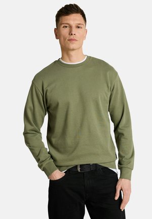 Mann trägt olivgrünen Langarmpullover und schwarze Hose mit einer Hand in der Tasche vor einem schlichten Hintergrund.
