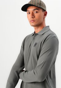 Olivenfarbener Polohemd mit langen Ärmeln aus strukturiertem Material und einem kleinen Logopatch. Kombiniert mit einer gemusterten Kappe mit schwarzem Schirm.
