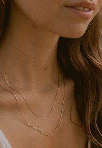 Roségoldene Halsketten in unterschiedlichen Längen übereinander getragen; eine ist strukturiert, während die andere glatt ist. Um den Hals getragen, komplementiert sie den Hautton.