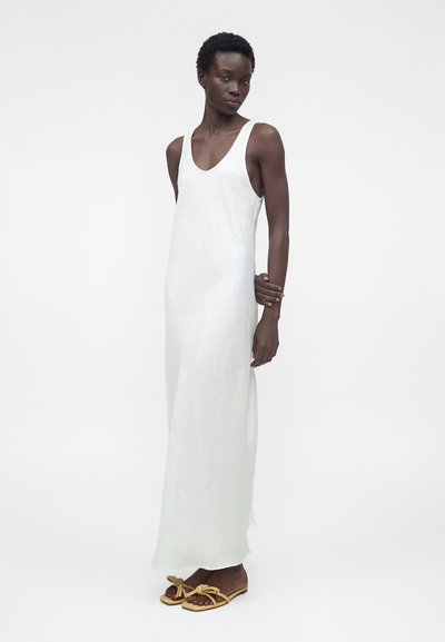 Γυναίκα με κοντά μαλλιά που φοράει ένα λευκό μακρύ φόρεμα χωρίς μανίκια και μπεζ επίπεδα σανδάλια με κόμπο, στέκεται μπροστά σε λευκό απλό φόντο.