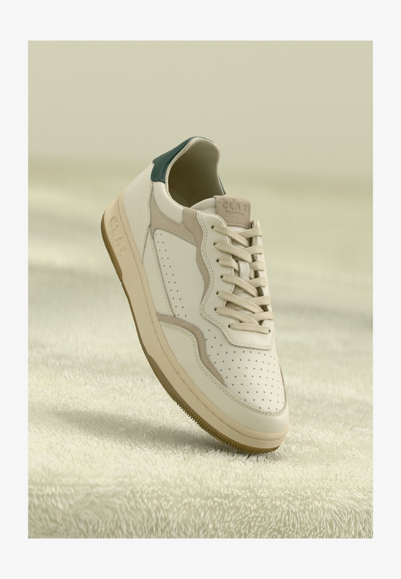Balti ir smėlio spalvos odiniai sportbačiai su perforacijomis, smėlio spalvos raišteliais, žaliu kulno elementu ir guminiu padu, pakabinti virš minkštos tekstūruotos paviršiaus.