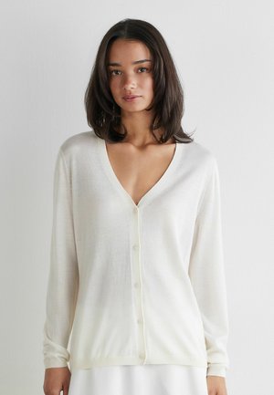 Ung kvinde med skulderlængde mørkt hår iført en hvid V-hals cardigan med knapper over en hvid nederdel mod en ensfarvet baggrund.