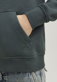 Dettaglio di una felpa verde scuro che mostra una mano in tasca, polsini a costine e tessuto testurizzato, abbinata a un paio di jeans in denim sbiaditi.