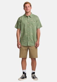 Camicetta a maniche corte di colore verde chiaro con motivi sottili, abbinata a pantaloni corti beige e scarpe da ginnastica nere, accompagnata da calze bianche.