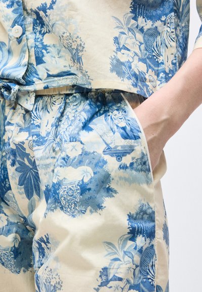 Μπλε και άσπρο πουκάμισο με λουλουδάτο μοτίβο και παντελόνι με μοτίβα λιονταριού και βάζου, το χέρι ακουμπάει στην τσέπη του παντελονιού.