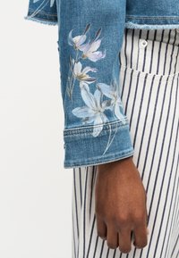 Μανίκι τζιν μπουφάν με λευκό και μοβ φλοράλ σχέδιο φορεμένο πάνω από το χέρι και ριγέ λευκό και μπλε παντελόνι.