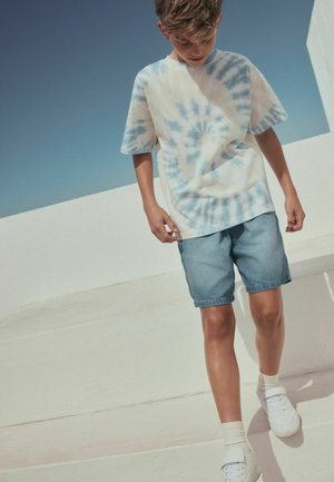 Băiat care merge afară purtând un tricou tie-dye albastru deschis, pantaloni scurți din denim, șosete albe și adidași albi, pe fundalul unui cer senin.