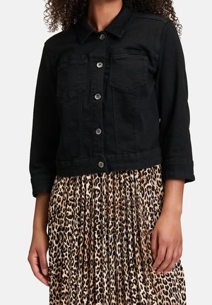 Kvinde iført en sort denimjakke med knapper over et plisseret nederdel med leopardtryk, stående foran en ensfarvet hvid baggrund.