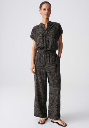 Frau trägt ein schwarz und dunkelgrün gemustertes kurzärmliges Hemd mit Knöpfen und passende Hosen mit weitem Bein sowie weiße Flip-Flops.