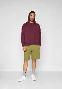 Mann in einem bordeauxfarbenen Reißverschluss-Hoodie mit aufgesetzter Kapuze, passendem T-Shirt, olivgrünen Shorts, grauen Socken und weißen Sneakers, der vor einem weißen Hintergrund steht.