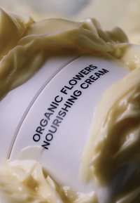 Vit burk märkt "Ekologisk blommor närande kräm", omgiven av en mjuk, krämig, ljusgul konsistens.