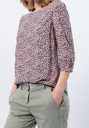 Femme portant un chemisier à motifs multicolores avec des manches trois-quarts et un pantalon gris clair, les mains dans les poches.