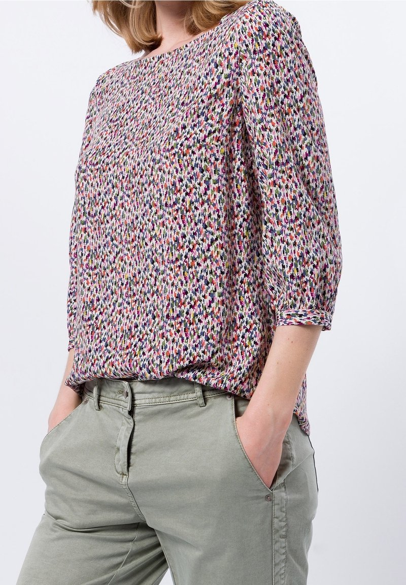 Femme portant un chemisier à motifs multicolores avec des manches trois-quarts et un pantalon gris clair, les mains dans les poches.