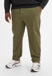 Olivgrüne Chinos mit geradem Schnitt, gefertigt aus weichem Baumwollstoff. Getragen mit weiß-silbernen Sneakers für einen lässigen Stil.
