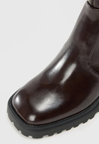 Braune Leder-Ankle-Boots mit glänzender Oberfläche, quadrater Zehenform und robustem schwarzen Gummisohle, die auffällige Profil- und Nähdetails aufweist.
