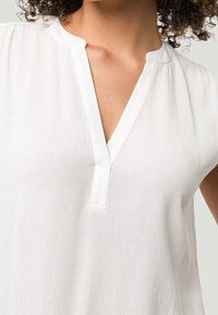 Blusa blanca con escote en V, mangas cortas y un acabado texturizado. La tela ligera resalta el diseño suave y fluido, así como las costuras sutiles.