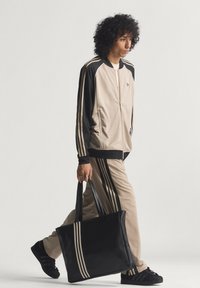 Beež ja musta värvi spordijakk kolme triibuga, sobivad püksid ning must kandekott. Lokkis juuksed, hoiab kotti ühe käega.