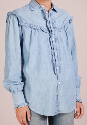 Hellblaues, langärmeliges Jeanshemd mit Rüschenbesatz entlang der Knopfleiste, am Rückenpasse, an den Manschetten und Schultern, getragen mit hell waschgewaschenen Jeans.